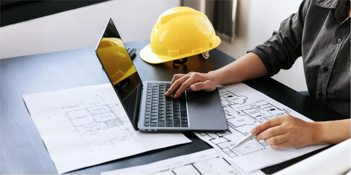 A person engrossed in work on a laptop, surrounded by architectural blueprints and a yellow hard hat on the desk, seamlessly integrates fluid system spreadsheets for engineering precision.