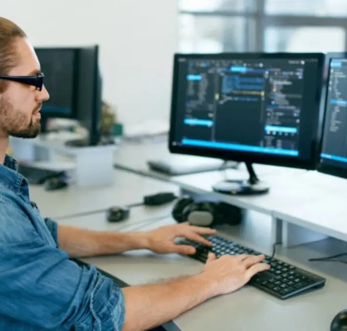 A person at a desk wearing a blue denim shirt and glasses uses dual monitors displaying PLM low-code solutions on a computer.
